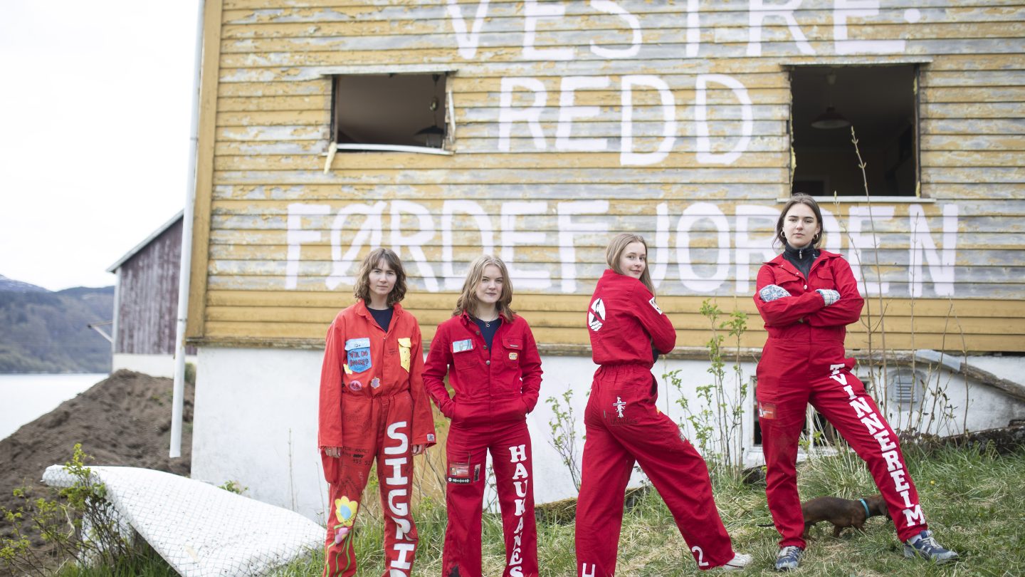 Fire jenter i russedress som står på plenen foran et gult hus med teksten Vestre redd førdefjorden